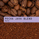 Coffee photograph showing whole roasted coffee beans at the top, roasted coffee grounds at the bottom, and a lavender label across the center reading “Mocha java Blend. Dark”.