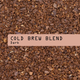 A close-up photo of dark brown cold brew coffee grounds with a horizontal muted peach label across the center reading 'COLD BREW BLEND. Dark'.