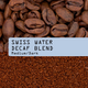 Coffee blend product photo showing whole roasted coffee beans at the top, a light blue label reading 'SWISS WATER DECAF BLEND Medium/Dark' in the center, and fine coffee granules at the bottom.