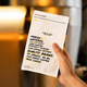 A hand holding a white coffee bag package with bold black and yellow text in a warm, blurred indoor setting.