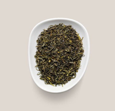 Dried loose-leaf tea leaves in a white oval dish, photographed from above on a light beige background.