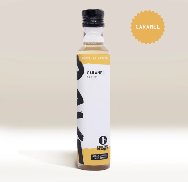 A bottle of caramel syrup with a black cap and a white label featuring black brushstroke graphics and yellow accents. A yellow starburst badge in the top right reads 'CARAMEL'.